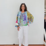 Woman wearing a colorful floral jacket and white pants standing against a white wall.