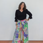 Woman wearing a black top and colorful patterned pants against a white wall.