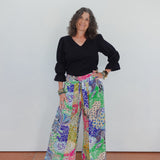 Woman wearing a black top and colorful patterned pants against a white wall.