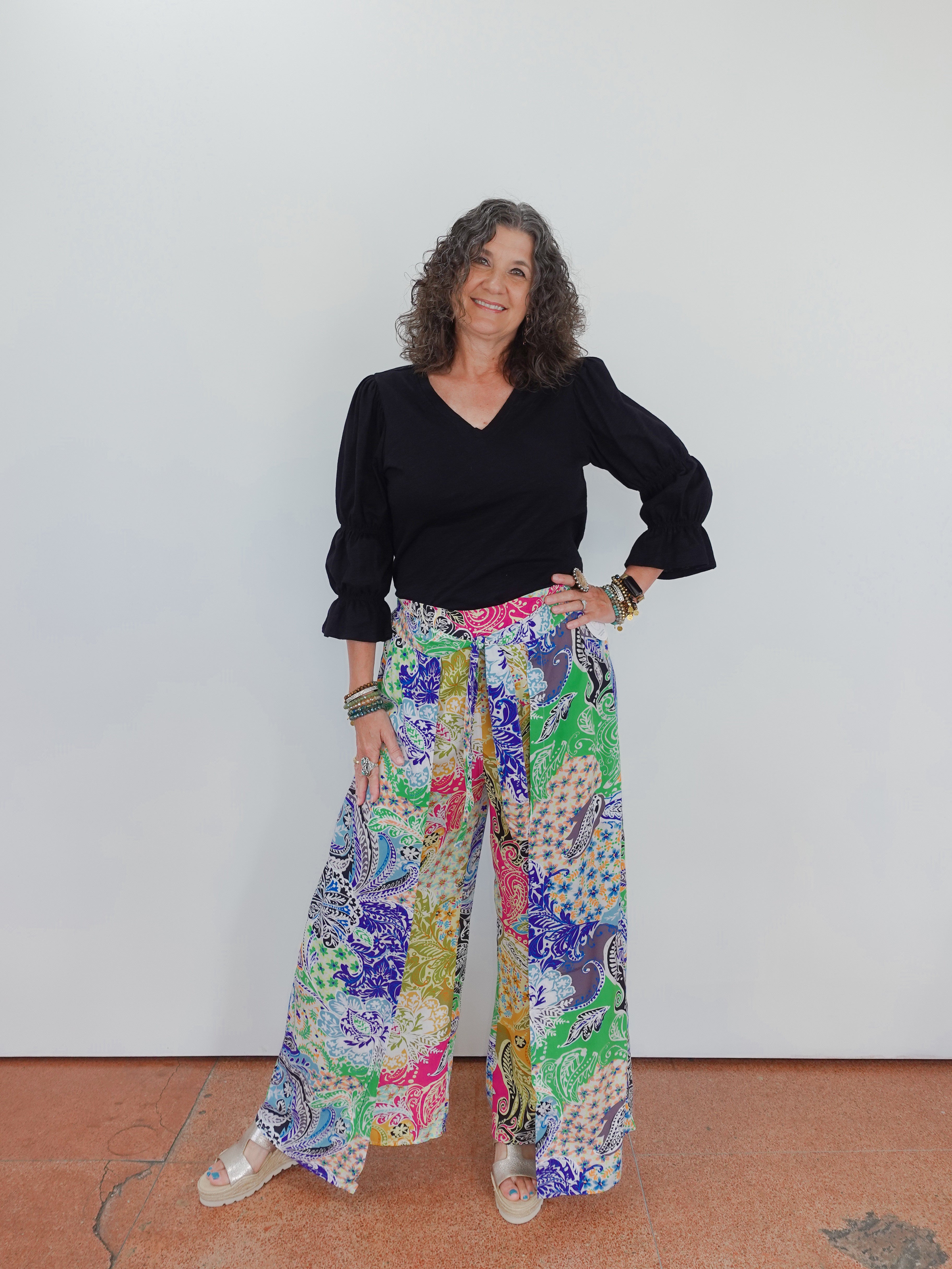 Woman wearing a black top and colorful patterned pants against a white wall.