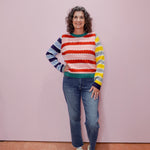 Woman wearing a colorful striped sweater and jeans standing against a pink wall.