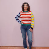 Woman wearing a colorful striped sweater and jeans standing against a pink wall.