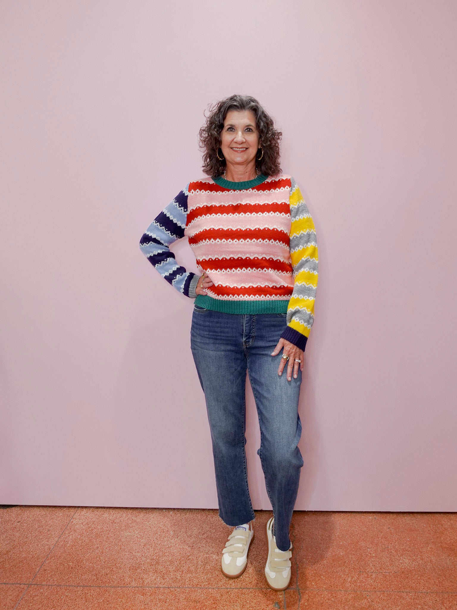 Woman wearing a colorful striped sweater and jeans standing against a pink wall.