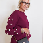 Woman wearing a burgundy outfit with pearl details and holding a black handbag against a white background