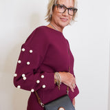 Woman wearing a burgundy outfit with pearl details and holding a black handbag against a white background