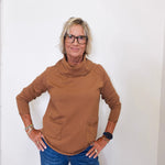 Casual outfit featuring a tan cowl neck sweater and blue jeans on a woman by a white wall.