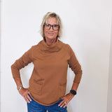 Casual outfit featuring a tan cowl neck sweater and blue jeans on a woman by a white wall.