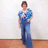 Woman wearing a colorful floral top and blue jeans standing against a white wall.