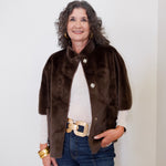 Woman wearing a brown fur vest over a white shirt with blue jeans against a plain background