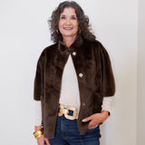 Woman wearing a brown fur vest over a white shirt with blue jeans against a plain background