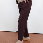 A person wearing dark brown straight-leg trousers, a textured beige and brown jacket, and beige woven wedge sandals, standing against a white wall on a terracotta-colored floor.