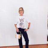 Woman wearing a graphic t-shirt with a cat design, dark jeans, and white sneakers against a white wall.