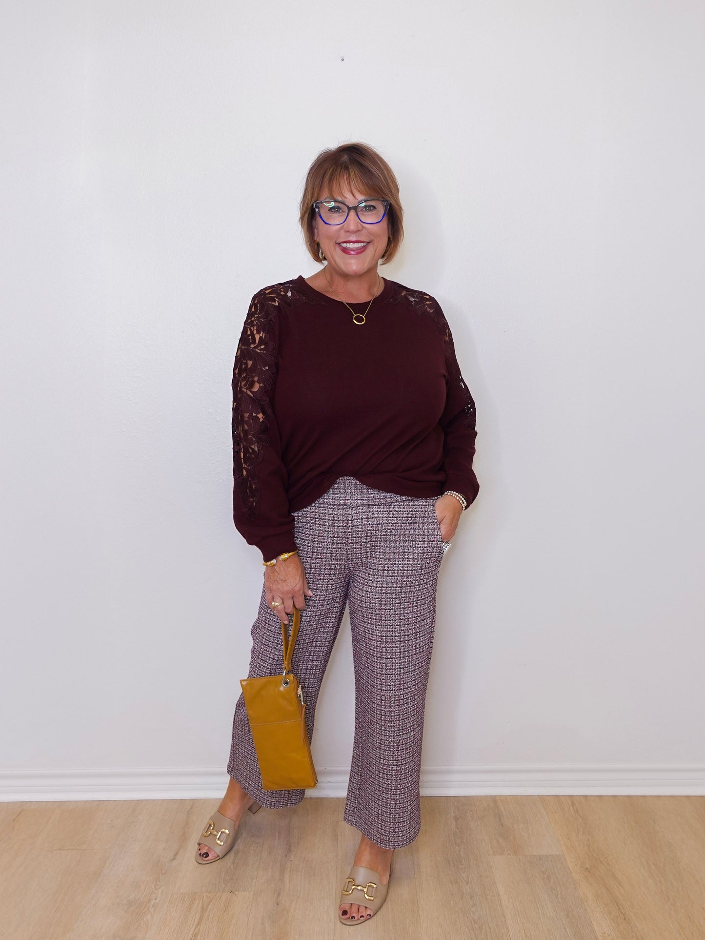 Woman wearing a burgundy sweater with lace sleeves and plaid pants, holding a yellow handbag against a white wall.