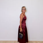 Woman in a burgundy dress holding a black handbag against a white wall.