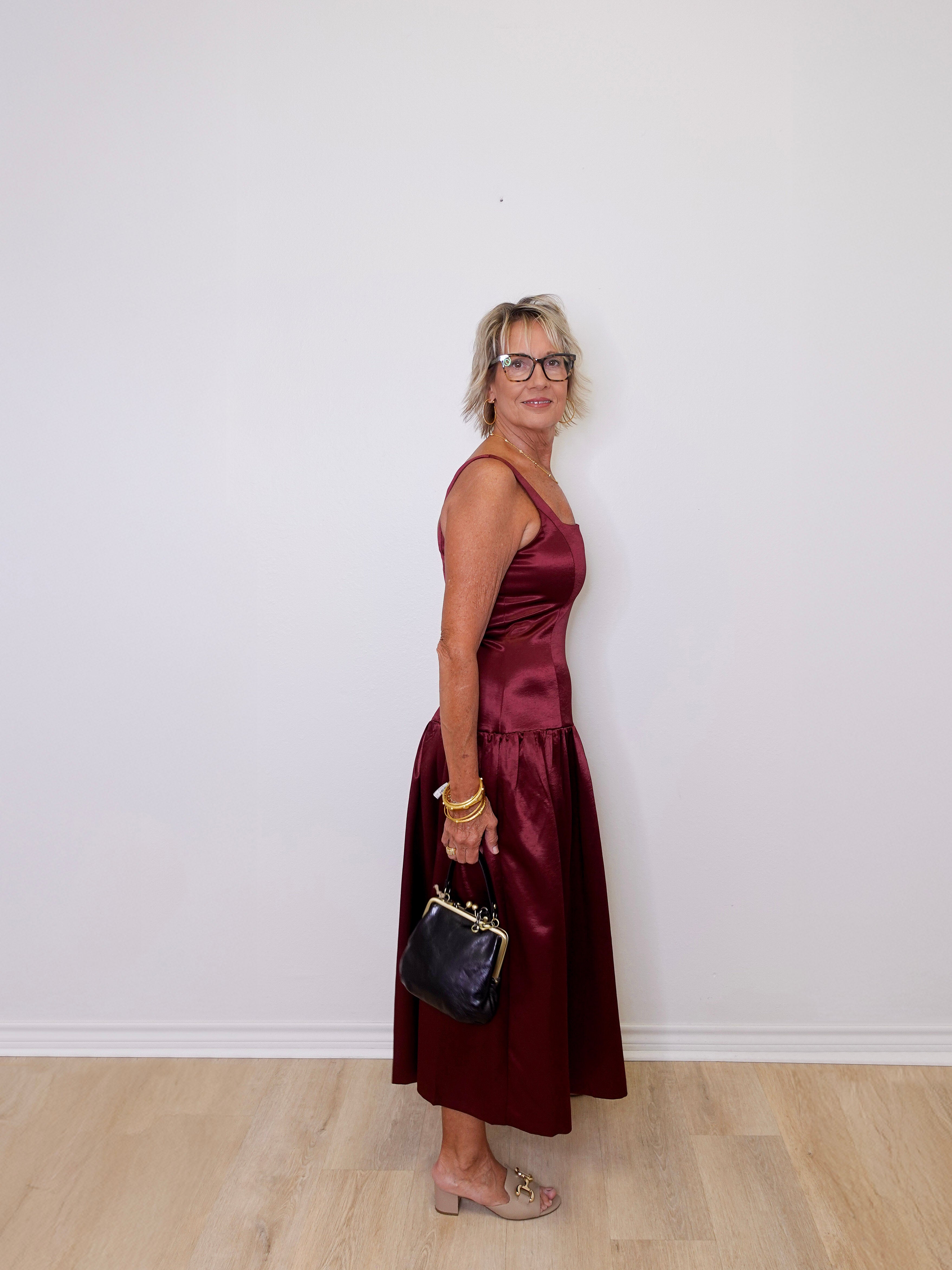 Woman in a burgundy dress holding a black handbag against a white wall.