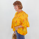 Woman wearing a yellow floral blouse and blue jeans, standing against a white wall.