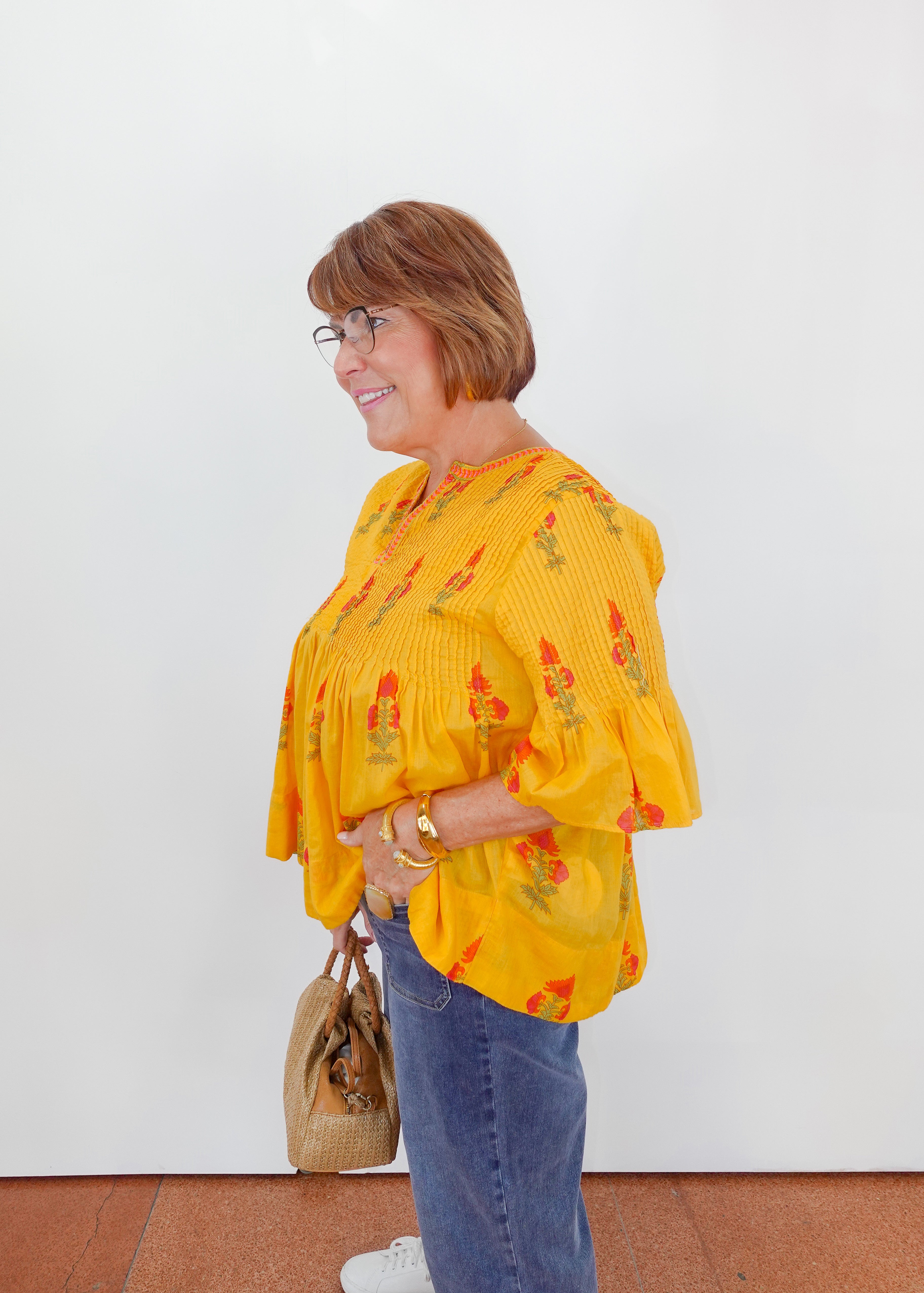 Woman wearing a yellow floral blouse and blue jeans, standing against a white wall.