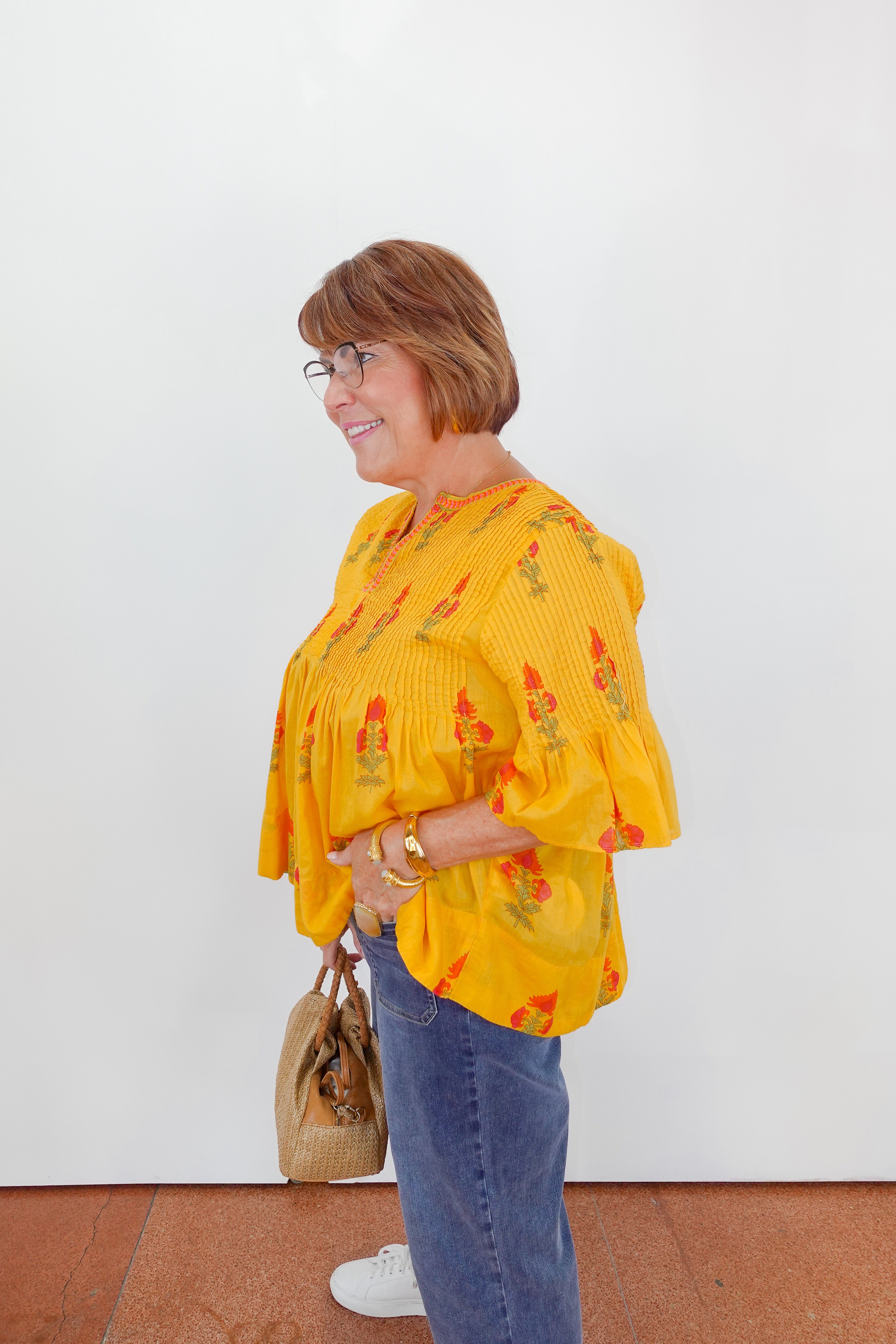 Woman wearing a yellow floral blouse and blue jeans, standing against a white wall.