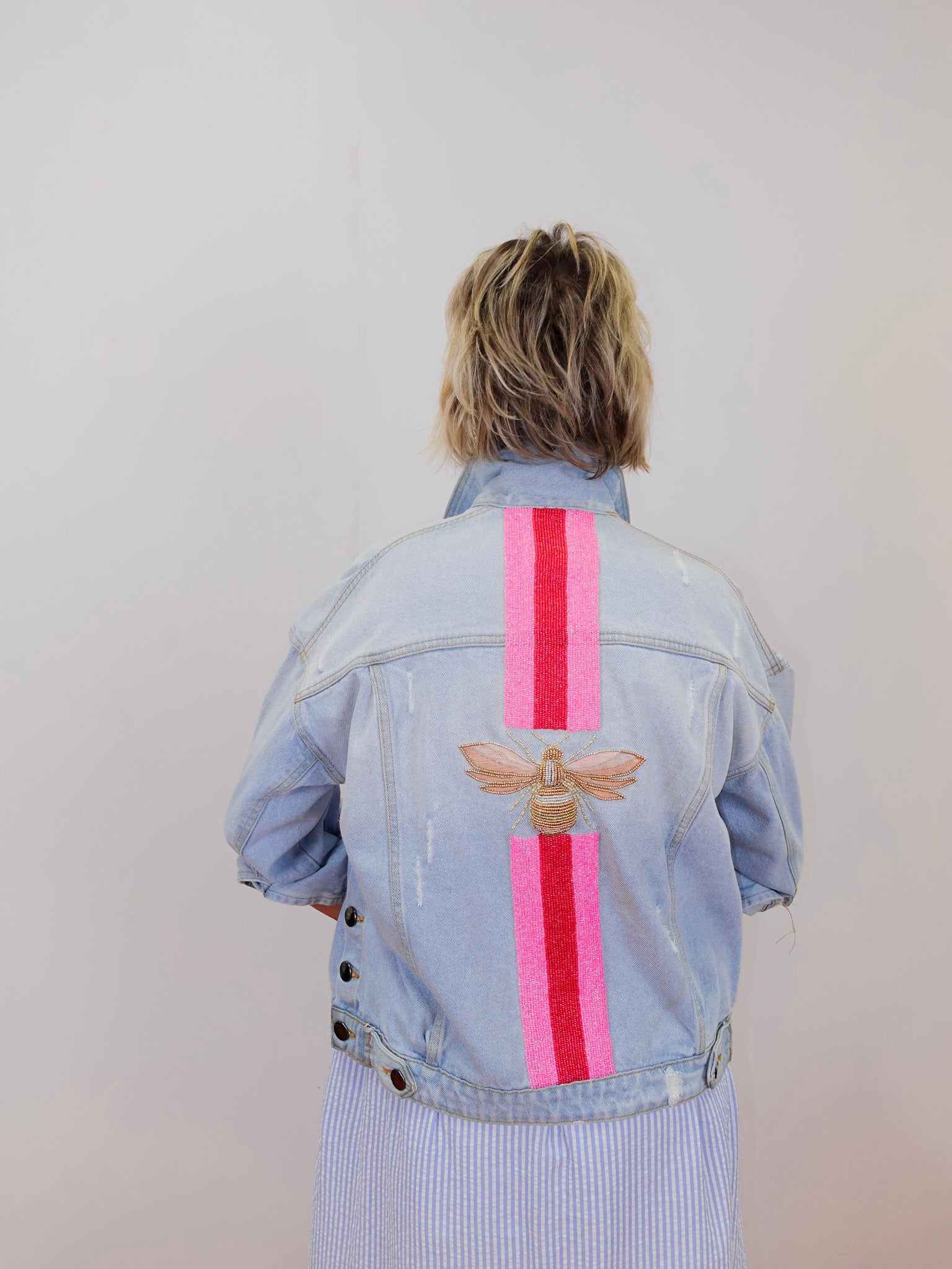 Person wearing a light blue denim jacket with pink stripes and a bee design on the back, standing against a plain background.