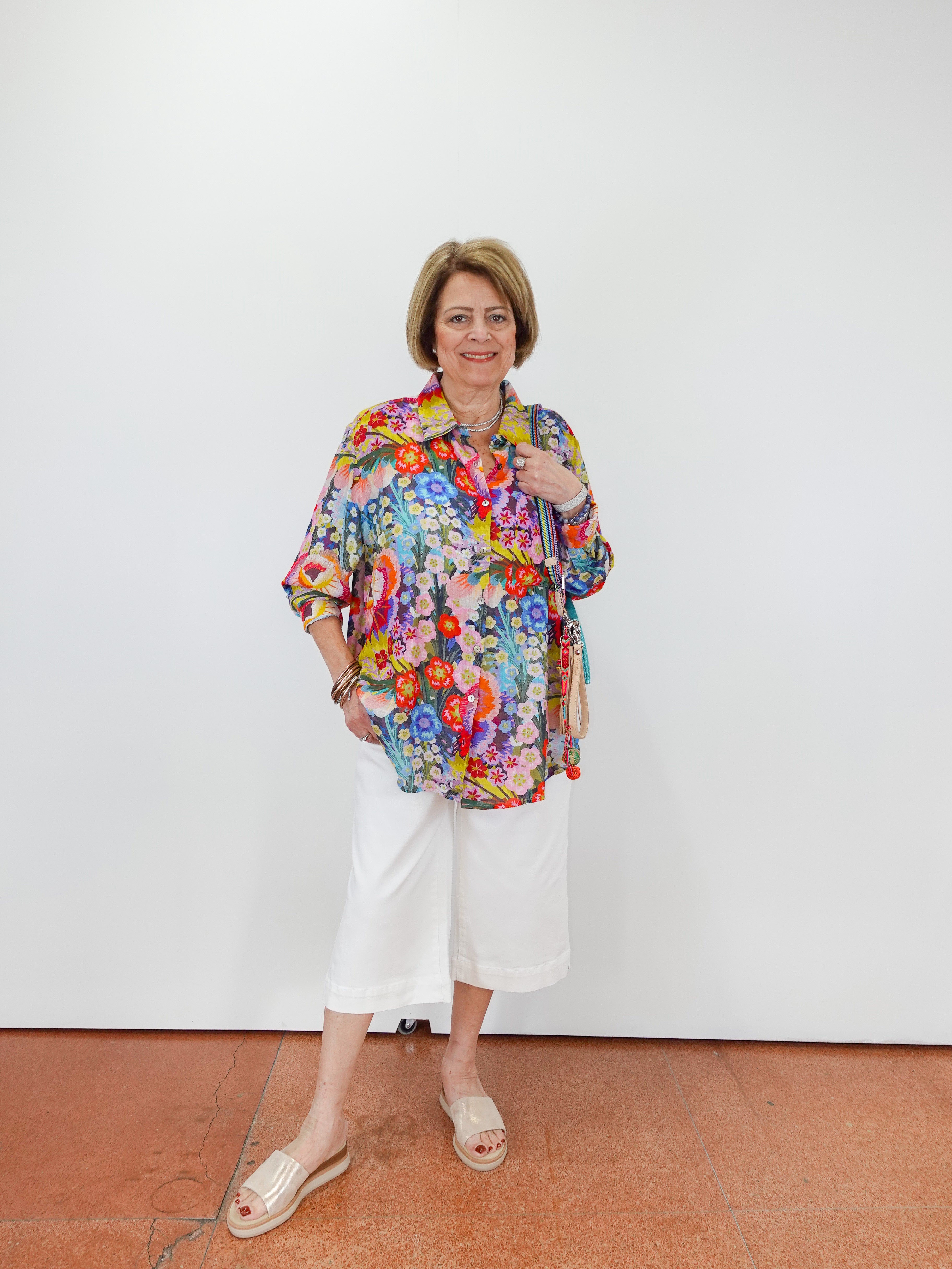 Woman wearing a colorful floral blouse and white pants against a white wall.