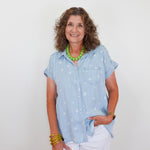 Woman wearing a light blue shirt with star patterns, white pants, and green necklace against a plain background