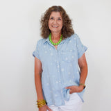 Woman wearing a light blue shirt with star patterns, white pants, and green necklace against a plain background