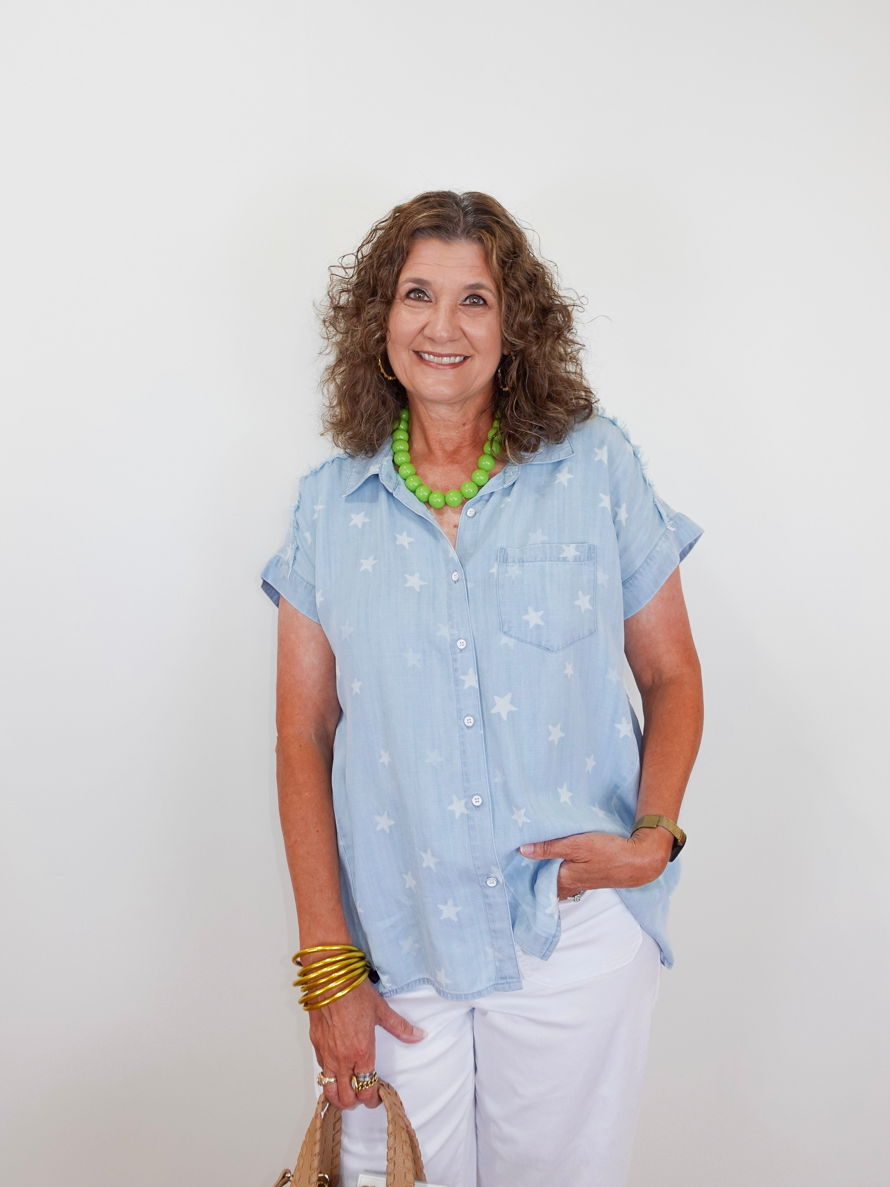 Woman wearing a light blue shirt with star patterns, white pants, and green necklace against a plain background