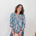 Woman wearing a colorful floral blouse and blue jeans, holding a beige clutch against a white wall.
