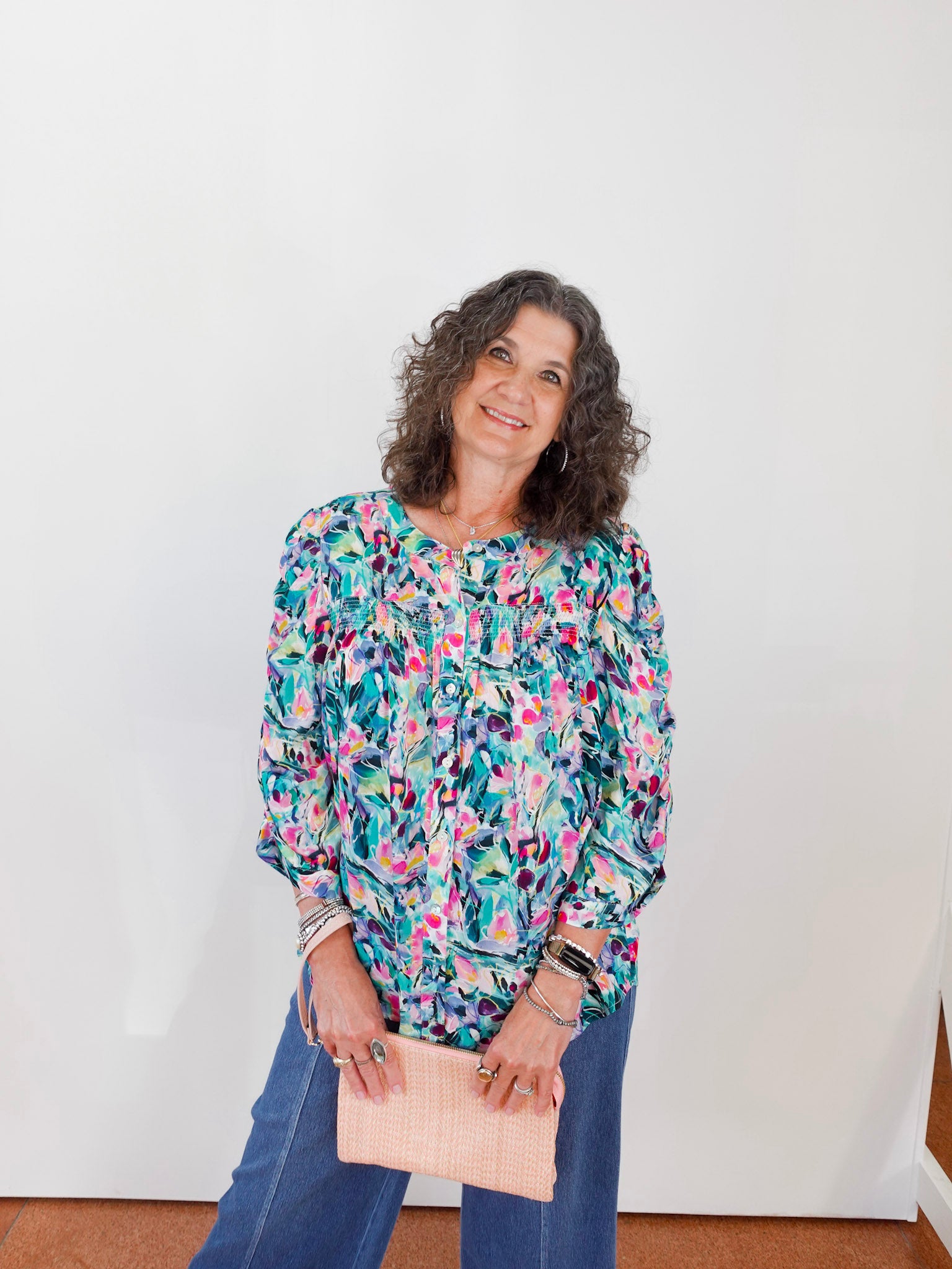 Woman wearing a colorful floral blouse and blue jeans, holding a beige clutch against a white wall.