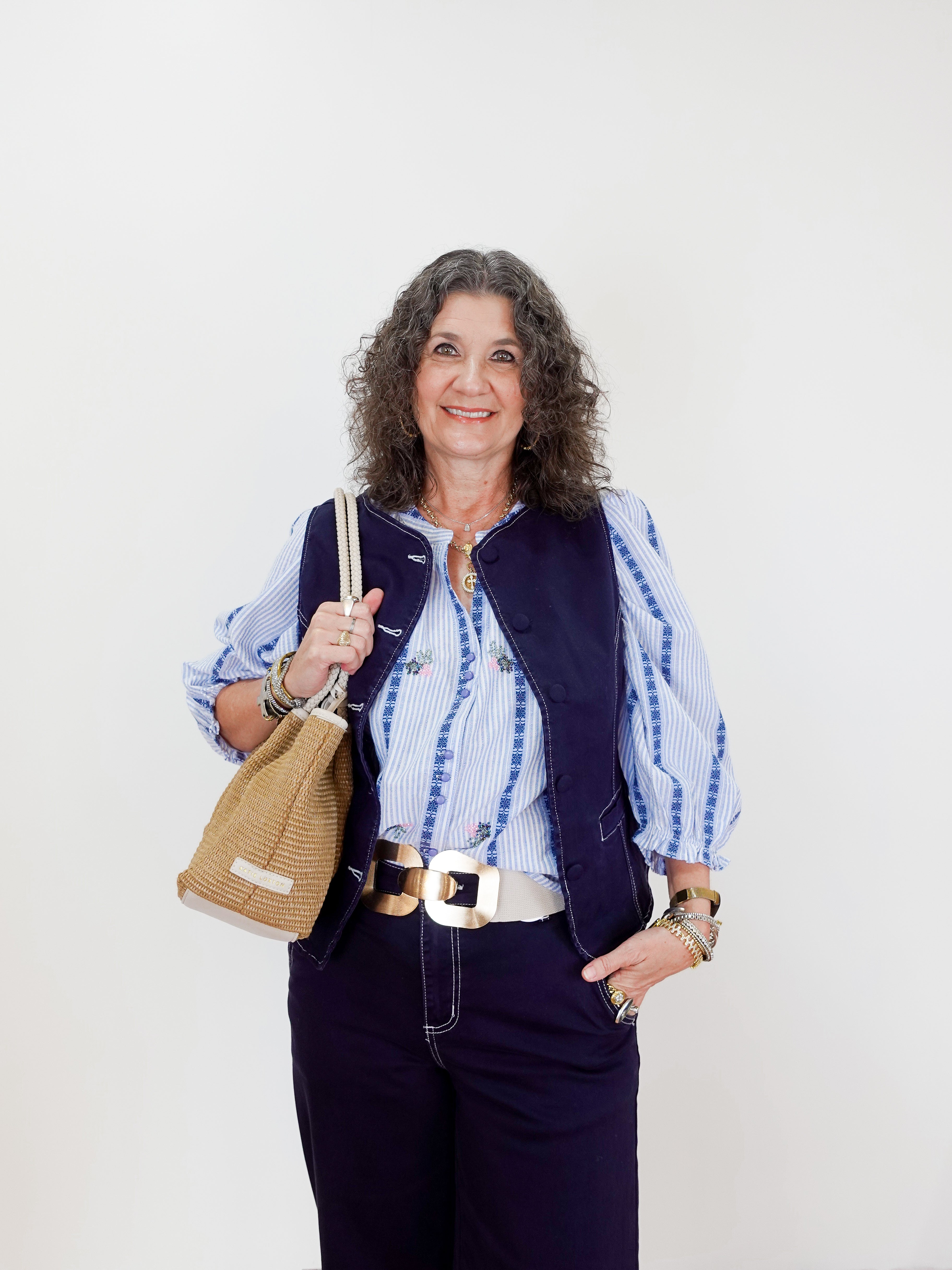 Woman wearing a blue vest over a striped shirt with dark pants, holding a woven bag against a white background.