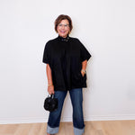 Woman wearing a black top and blue jeans standing against a white wall.