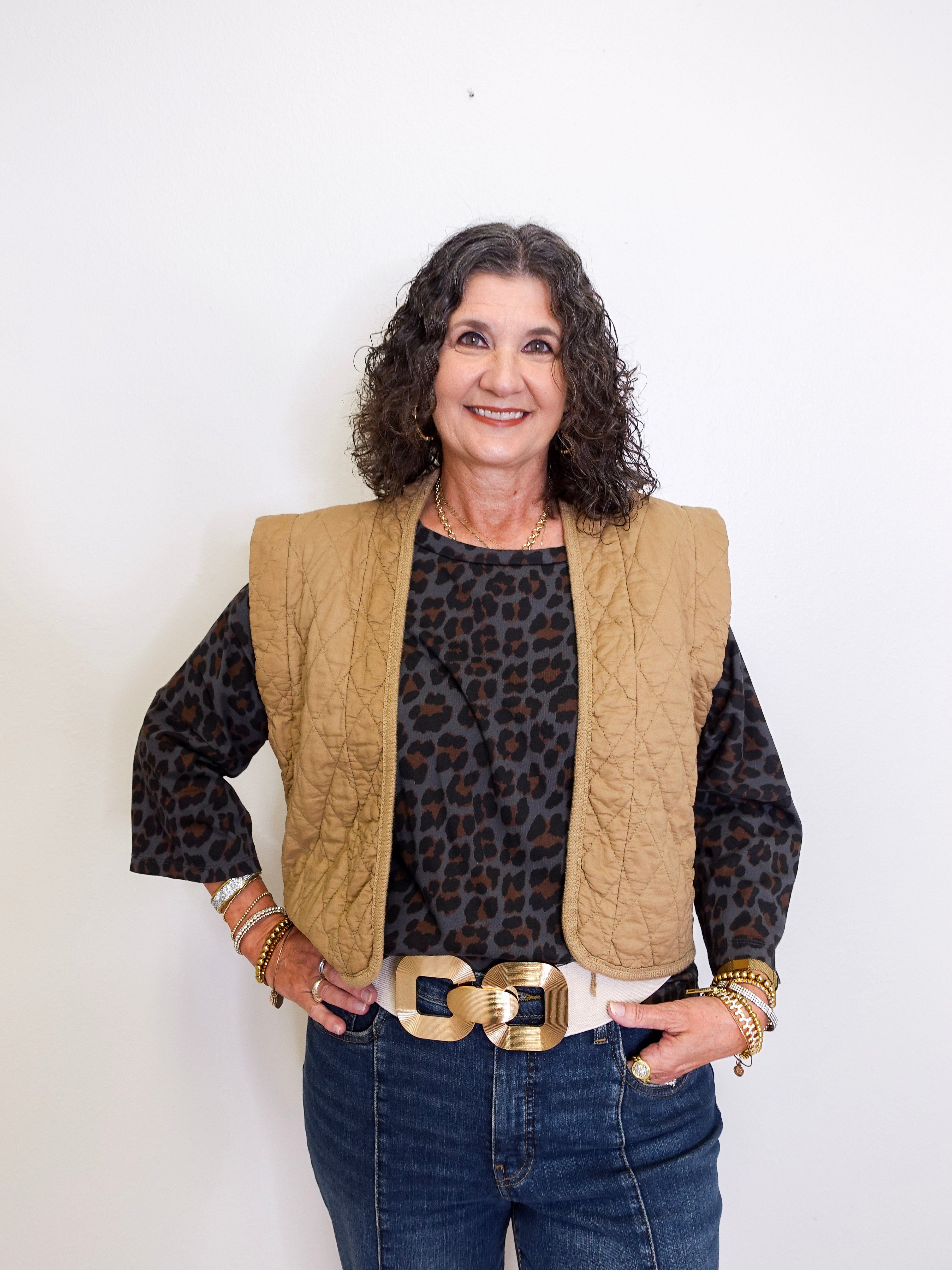 Woman wearing a leopard print top, beige vest, and blue jeans against a white background