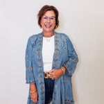 Woman wearing a blue embroidered jacket and denim jeans against a white wall