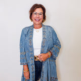 Woman wearing a blue embroidered jacket and denim jeans against a white wall