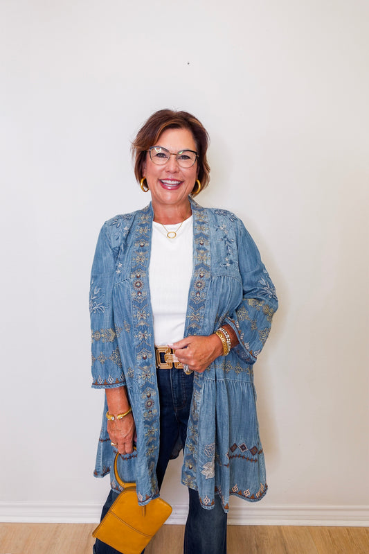 Woman wearing a blue embroidered jacket and denim jeans against a white wall