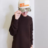 Person wearing a cap with 'HE'S HUNTING' text against a white wall