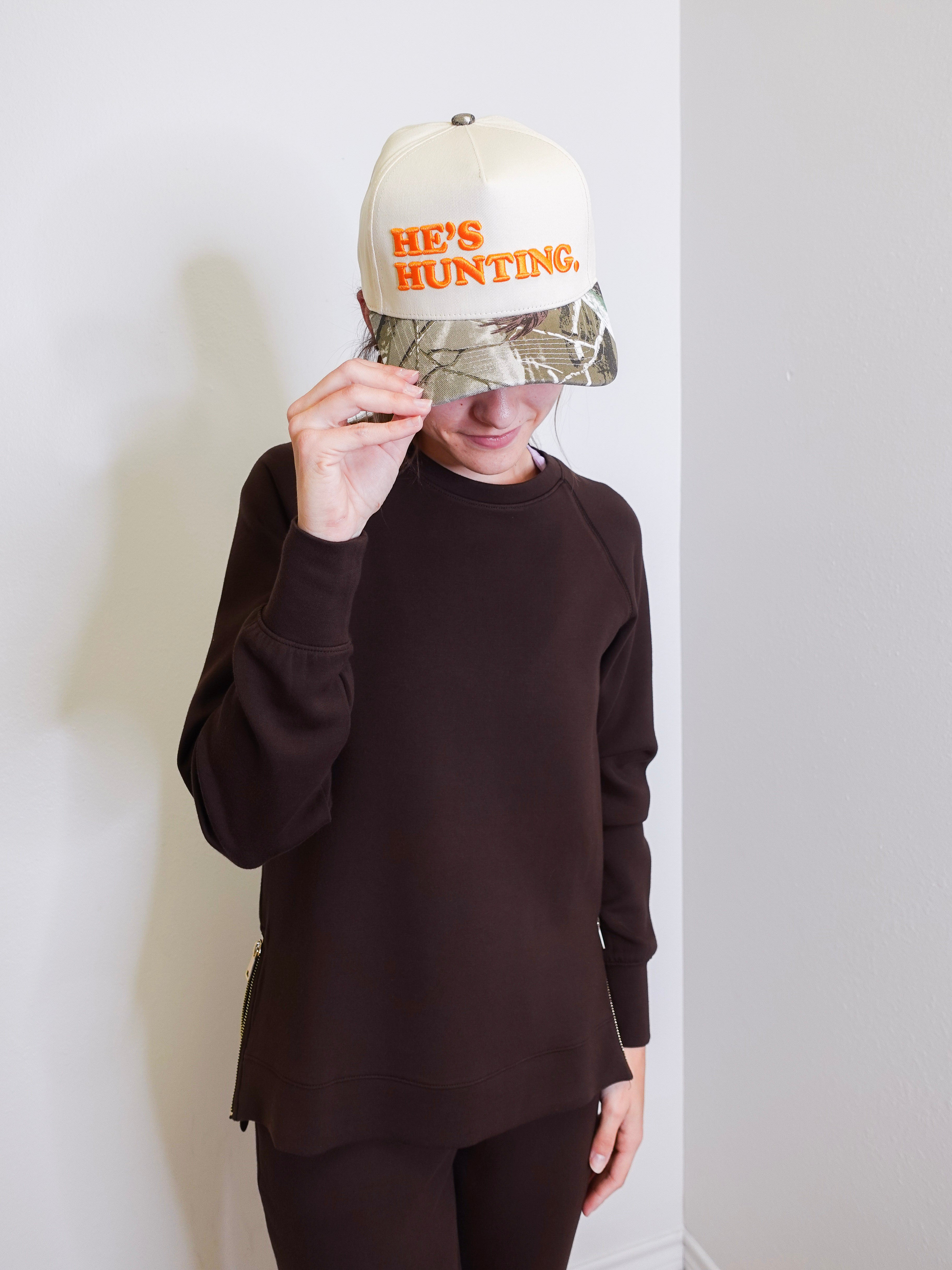 Person wearing a cap with 'HE'S HUNTING' text against a white wall