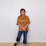 Woman wearing a brown jacket and blue jeans standing against a white wall.