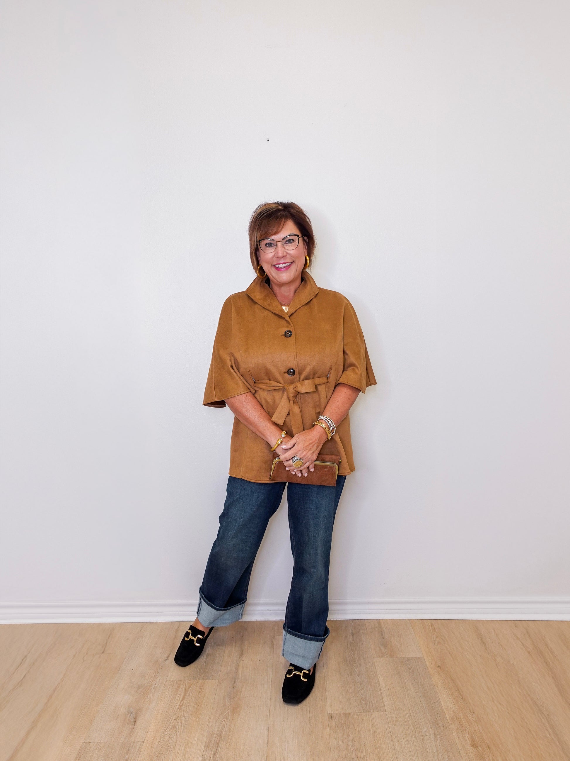 Woman wearing a brown jacket and blue jeans standing against a white wall.