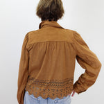 A person shown from the back wearing a brown suede-like long-sleeve blouse with a scalloped eyelet lace hem, paired with light blue jeans, against a plain light gray background.