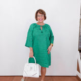 Woman in a green dress holding a white handbag against a white wall.