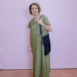 Woman in a green dress with floral patterns standing against a light purple wall.