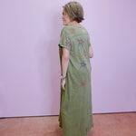 Woman wearing a green dress with floral patterns standing against a plain wall.