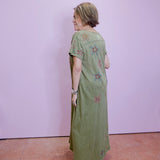 Woman wearing a green dress with floral patterns standing against a plain wall.