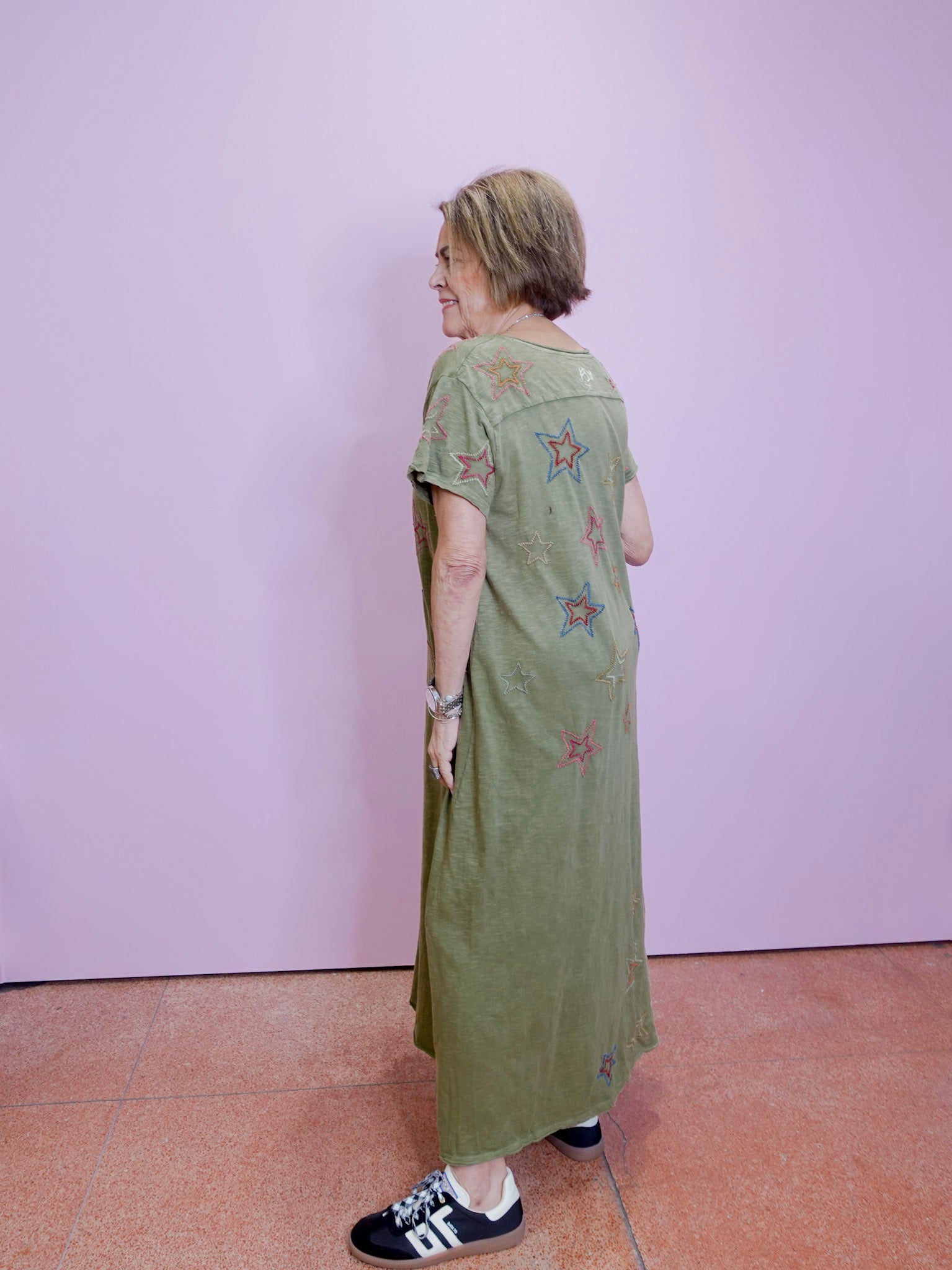 Woman wearing a green dress with floral patterns standing against a plain wall.