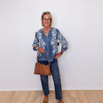 Woman wearing a blue floral blouse, jeans, and brown shoes against a white wall.