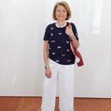Woman wearing a navy sweater with pink patterns and white pants, holding a red bag against a white wall.