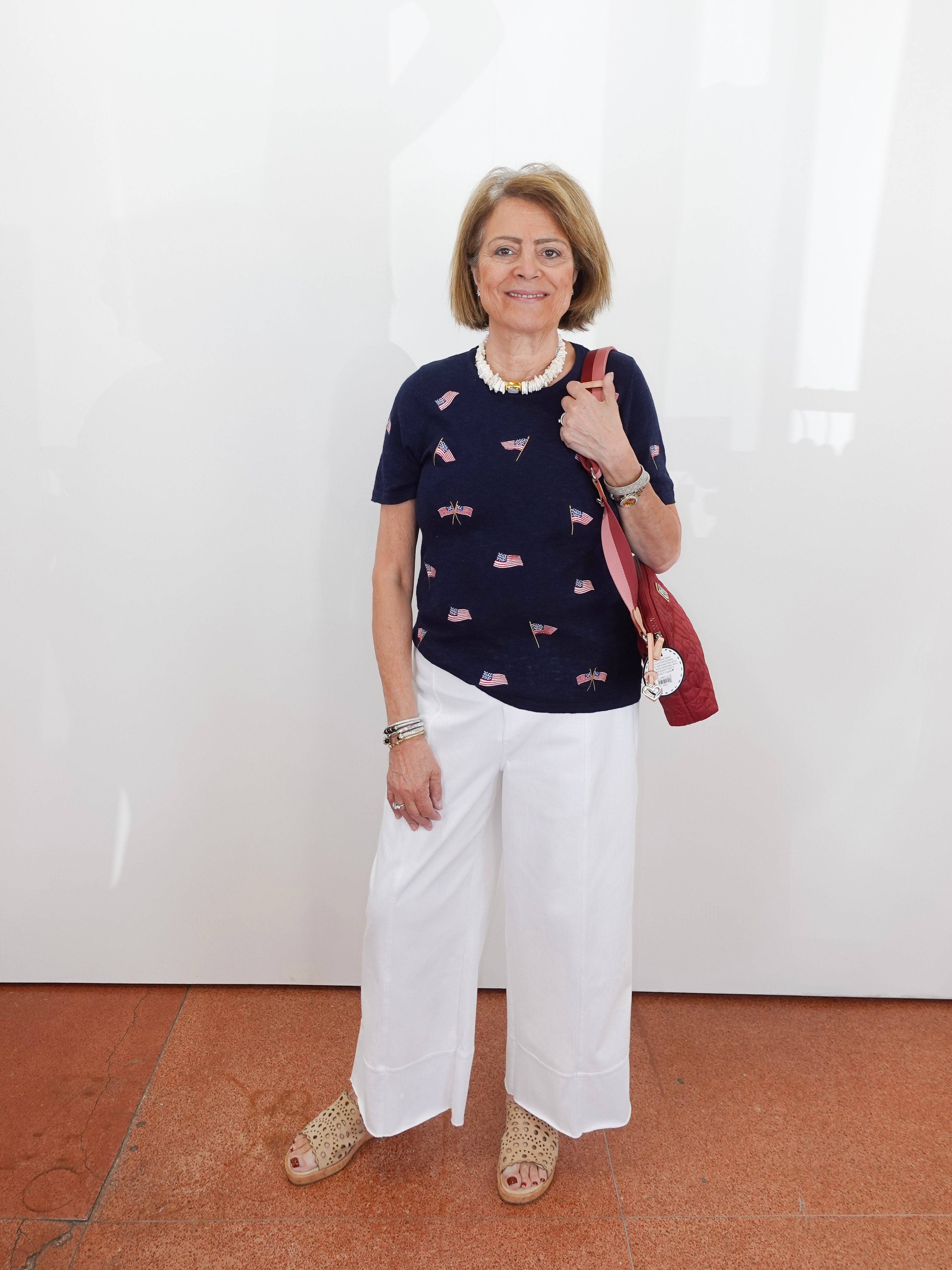 Woman wearing a navy sweater with pink patterns and white pants, holding a red bag against a white wall.