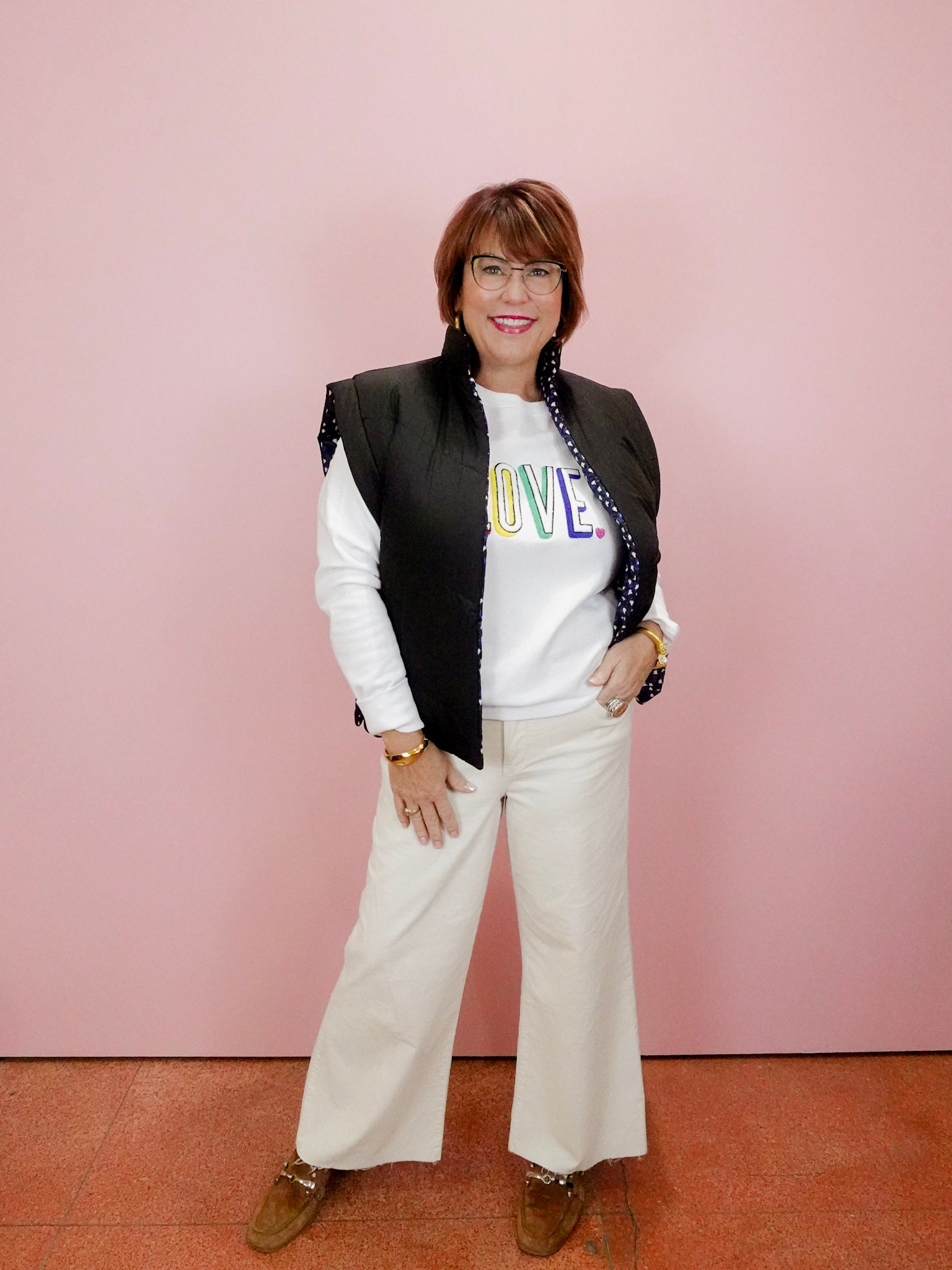 Woman wearing a black vest over a white shirt with colorful text, standing against a pink wall.