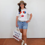 Woman wearing a white t-shirt with colorful design, denim shorts, and a hat, holding a tote bag.